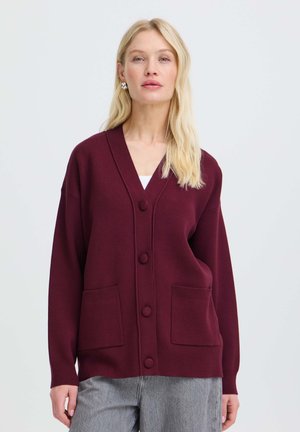Donna con capelli biondi lunghi indossa un cardigan bordeaux abbottonato con due tasche frontali sopra una maglia bianca e pantaloni grigi su uno sfondo semplice.