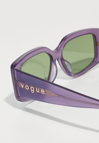 Μωβ μεγάλες ηλιό γυαλιά με πράσινους φακούς. Διαθέτουν χοντρό, διαφανές σκελετό και μεταλλικό λογότυπο "vogue" στο πλάι.