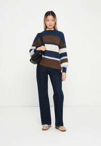 Pull rayé marron, marine et crème avec une coupe décontractée ; associé à un jean bleu foncé et des chaussures beiges. Modèle portant un sac noir.