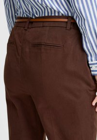 Braune Hose aus einem glatten Stoff mit Gesäßtaschen und flacher Vorderseite, kombiniert mit einem blau-weiß gestreiften Hemd und einem braunen Gürtel.