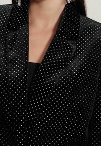 Blazer en velours noir à pois argentés, avec des revers crantés et une texture lisse. Mise au point sur la moitié supérieure, dévoilant en partie un haut noir en dessous.