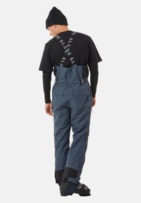 Blaue Skihosen mit verstellbaren Trägern, die ein schlankes Design, Reißverschlusstaschen und verstärkte schwarze Bündchen aufweisen. Getragen mit einer schwarzen Mütze.