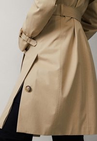 Beżowy płaszcz trench z wiązanym w pasie, długimi rękawami i guzikami na dole. Wykonany z gładkiego, lekkiego materiału o dopasowanym kroju.