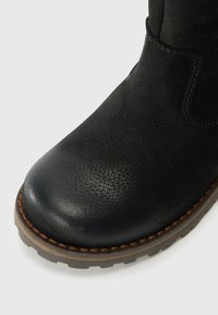Botte en cuir noir avec une finition texturée, bout rond et une semelle en caoutchouc robuste. Présente un détail de couture marron le long du bord de la semelle.