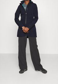 Giacca impermeabile blu navy con cerniera frontale, cappuccio regolabile e tessuto traspirante. Abbinata a pantaloni neri larghi e scarpe nere.