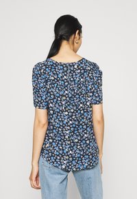 Blusa floreale nera con motivi blu, bianchi e verdi. Maniche corte a sbuffo e dettaglio a chiave sul retro. Abbinata a jeans azzurri chiari.