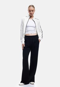 Witte zip-up bomberjack met zwarte strepen, gecombineerd met losse zwarte joggingbroeken. Model staat met één hand in de zak, met ronde oorbellen.