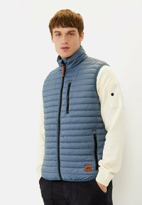 Hellblaues gesteppte Weste mit Stehkragen, Vorderreißverschluss-Taschen und braunen Lederelementen. Getragen über einem cremefarbenen Pullover, der Kragen ist sichtbar.