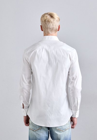 Chemise blanche à manches longues avec un col boutonné, présentant une texture lisse et un ourlet incurvé. Portée avec un jean déchiré bleu clair.