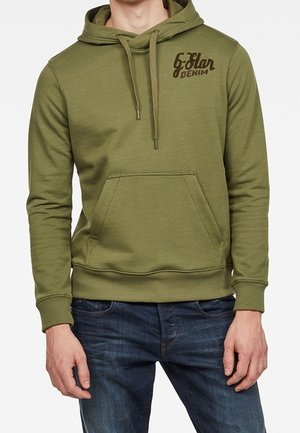 Homme portant un sweat à capuche vert olive avec poche avant et logo "G Star Denim" sur le haut à gauche de la poitrine, associé à un jean bleu.