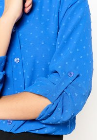 Camicia azzurra con bottoni, caratterizzata da un motivo tessurato. Presenta maniche arrotolabili, piccoli bottoni blu e un tessuto a trama liscia.