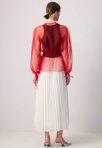 Durchsichtige rote Bluse mit Puffärmeln, tailliert, kombiniert mit einem plissierten weißen Rock und silberfarbenen Schuhen. Modell von hinten gezeigt.