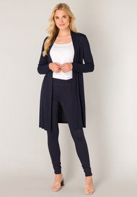 Marineblauwe lange cardigan gemaakt van zachte stof, gecombineerd met een wit tanktopje en marineblauwe legging, met een ontspannen pasvorm en een open voorkant.