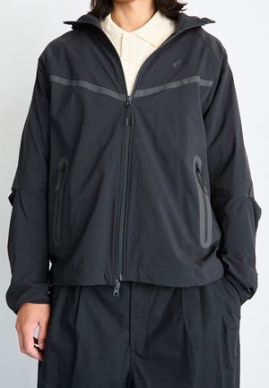 Personne portant une veste Nike noire zippée avec des bandes réfléchissantes horizontales sur une chemise beige à col, debout les mains dans les poches d'un pantalon noir.
