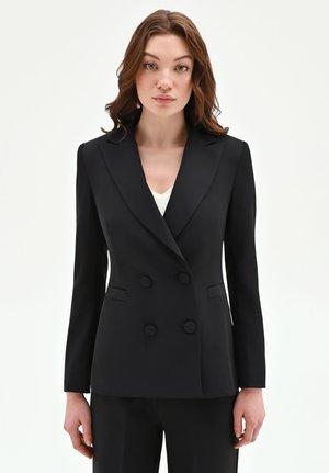 Donna dalla pelle chiara e capelli castani che indossa un blazer nero doppiopetto aderente e pantaloni neri, in piedi davanti a uno sfondo bianco.