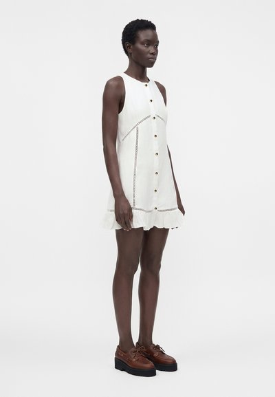 Femme debout portant une robe blanche sans manches boutonnée avec un ourlet à volants et des chaussures marron à plateforme à lacets sur fond uni.