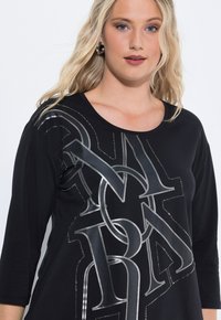 Schwarzes Oberteil mit drei Viertelärmeln, das ein metallisch silbernes Grafikdesign mit Buchstaben und gepunkteten Akzenten auf der Vorderseite präsentiert. Weicher, strukturierter Stoff.