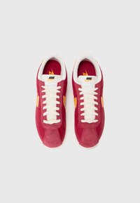 Baskets Nike rouges avec une tige en suède et en tissu, dotées de lacets blancs, d'accents jaunes et d'un intérieur rembourré.