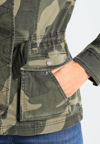 Zelené kárované sako se stahovacími šňůrkami v pase, cvokovými kapsami a prošívanou texturou na látce. Odolný materiál.