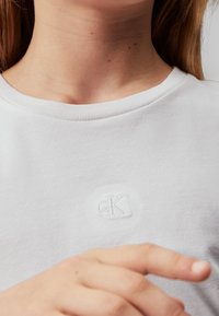 Biała bawełniana koszulka z okrągłym dekoltem i haftowanym logo "ck" w białym kolorze. Gładka faktura, krótkie rękawy, casualowy design.