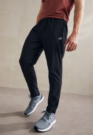 Mand iført sorte New Balance træningsbukser og grå New Balance sneakers står på en glat gulv mod en ensfarvet væg.