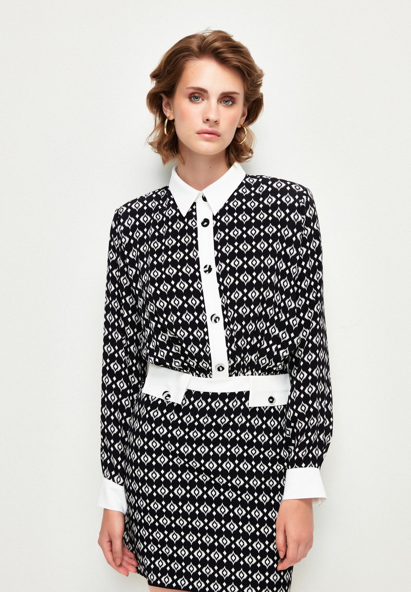 Schwarz-weiß gemustertes Kleid mit spitzen Kragen, langen Ärmeln, einem weißen Gürtel und Knopfdetails an der Vorderseite sowie an den Manschetten.