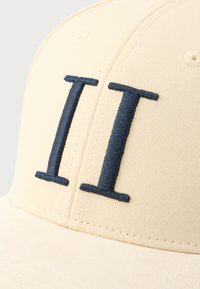 Beżowa czapka z daszkiem z widocznym granatowym haftowanym napisem "II" na przodzie, gładka w dotyku i z lekko zaokrąglonym daszkiem.