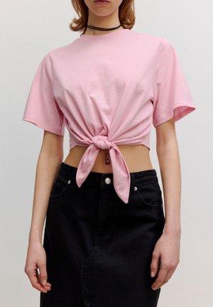 Kvinde iført en lyserød kort T-shirt bundet foran, kombineret med et sort højtaljet skørt og en synlig navlepiercing.