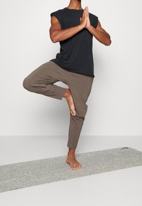 Sort ærmeløs skjorte og brune tapered bukser båret af en person i en yogastilling på en tekstureret grå måtte.