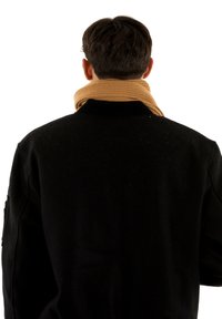 Uomo con capelli corti e scuri che indossa un maglione nero e una sciarpa marrone piegata, di spalle su uno sfondo bianco.