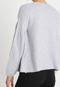 Vista trasera de una persona que lleva una camiseta de manga larga gris claro con mangas plisadas y pantalones ajustados negros.