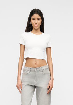 Hosszú sötét hajú nő, fehér, rövid ujjú, haspólót és világosszürke farmert visel, egyszínű háttér előtt áll.