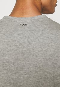 T-shirt din bumbac gri, cu guler rotund, având o textură fină. Brandingul negru "HUGO" se află în partea superioară a spatelui.
