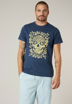 Man draagt een marineblauw T-shirt met een geel bloemen schedelontwerp, lichtblauwe korte broek, staat met ontspannen handen, neutrale achtergrond.