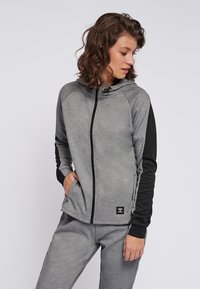 Sweat à capuche gris zippé avec un accent noir sur les manches, fabriqué à partir d'un tissu doux et texturé. Présente un col haut et des poches latérales.