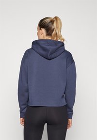 Mornarško modra skrajšana kapuco s kapuco, dolgimi rokavi in mehkobo. Kombinirana s tesnimi črnimi leggins, kar prikazuje sproščeno športno podobo.