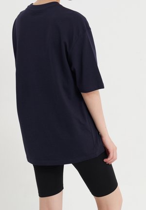 Person iført en løs mørkeblå t-shirt og tætsiddende sorte shorts, vist bagfra mod en ensfarvet lys baggrund.