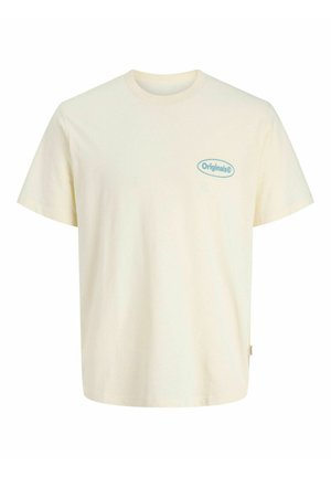 Ung person iført beige t-shirt med logo, mørke denimshorts, beige kasket, hvide sokker og brune snørestøvler står mod en lys baggrund.
