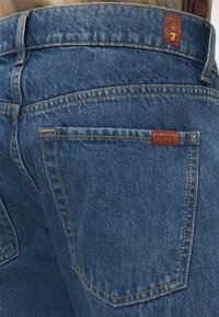 Περιοχή πίσω τσέπης σε μπλε τζιν denim με πορτοκαλί ραφές και δύο μικρές κόκκινες υφασμάτινες ετικέτες, εκ των οποίων η μία γράφει "for all mankind."