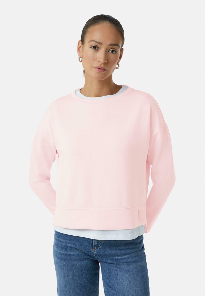 Lichtroze sweatshirt gemaakt van zacht materiaal, voorzien van een ronde halslijn, verlaagde schouders en een geribbelde rand, gecombineerd met een witte gelaagde shirt eronder.