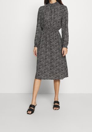 Femme portant une robe midi noire à petits motifs floraux blancs, manches longues, col haut, taille froncée, et sandales noires à bout ouvert.