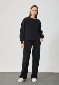 Vrouw staat gekleed in een losse zwarte sweatshirt, wijde zwarte broek en witte sneakers tegen een effen lichte achtergrond.