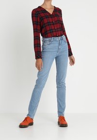 Chemise à carreaux rouge et noire avec boutons, jean bleu clair et chaussures orange. La tenue présente des couleurs contrastées et une coupe décontractée.