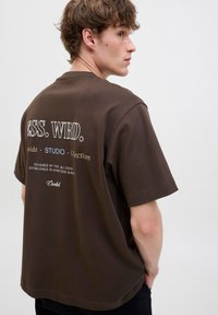 Brown cotton t-shirt featuring a crew neckline, short sleeves, and printed text on the back in white and blue. Accented with a relaxed fit.