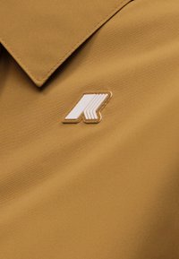 Κοντινό πλάνο σε μπεζ ύφασμα κολάρο με ένα λευκό και μπεζ επίπεδο γεωμετρικό σήμα με κονκάρδα, τοποθετημένο κοντά στη γραμμή του λαιμού.