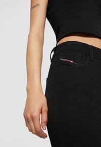 Black denim jeans with a fitted design. Features a small pocket with red and white branding. Smooth texture, visible stitching details.