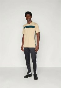 Beige atletisk t-shirt med mørkegrøn vandret stribe, parret med sorte bukser med hvide sidelister, iført sorte sneakers.