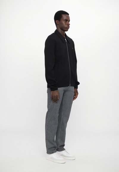 Veste zippée noire avec un col, associée à un jean gris et des baskets blanches. Le tissu semble lisse, avec un design simple et minimaliste.
