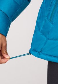 Une veste rembourrée bleu vif avec un design matelassé, dotée d'un cordon de serrage à l'ourlet pour l'ajustement, montrant un gros plan de la main qui le tire.