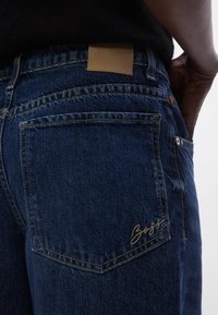 Kék farmernadrág sima textúrával, bőrpátával és hímzett logóval a hátizsebben, hagyományos varrást bemutatva.